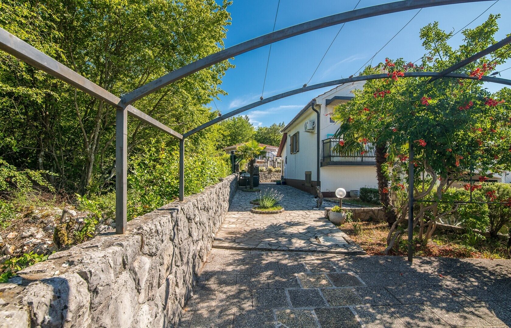 A stone pathway leading to a building with a white facade, surrounded by greenery and a metal pergola structure.