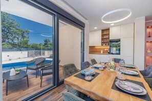 Uma sala de jantar com mesa de madeira posta para refeição, piscina externa e cozinha com armários brancos.
