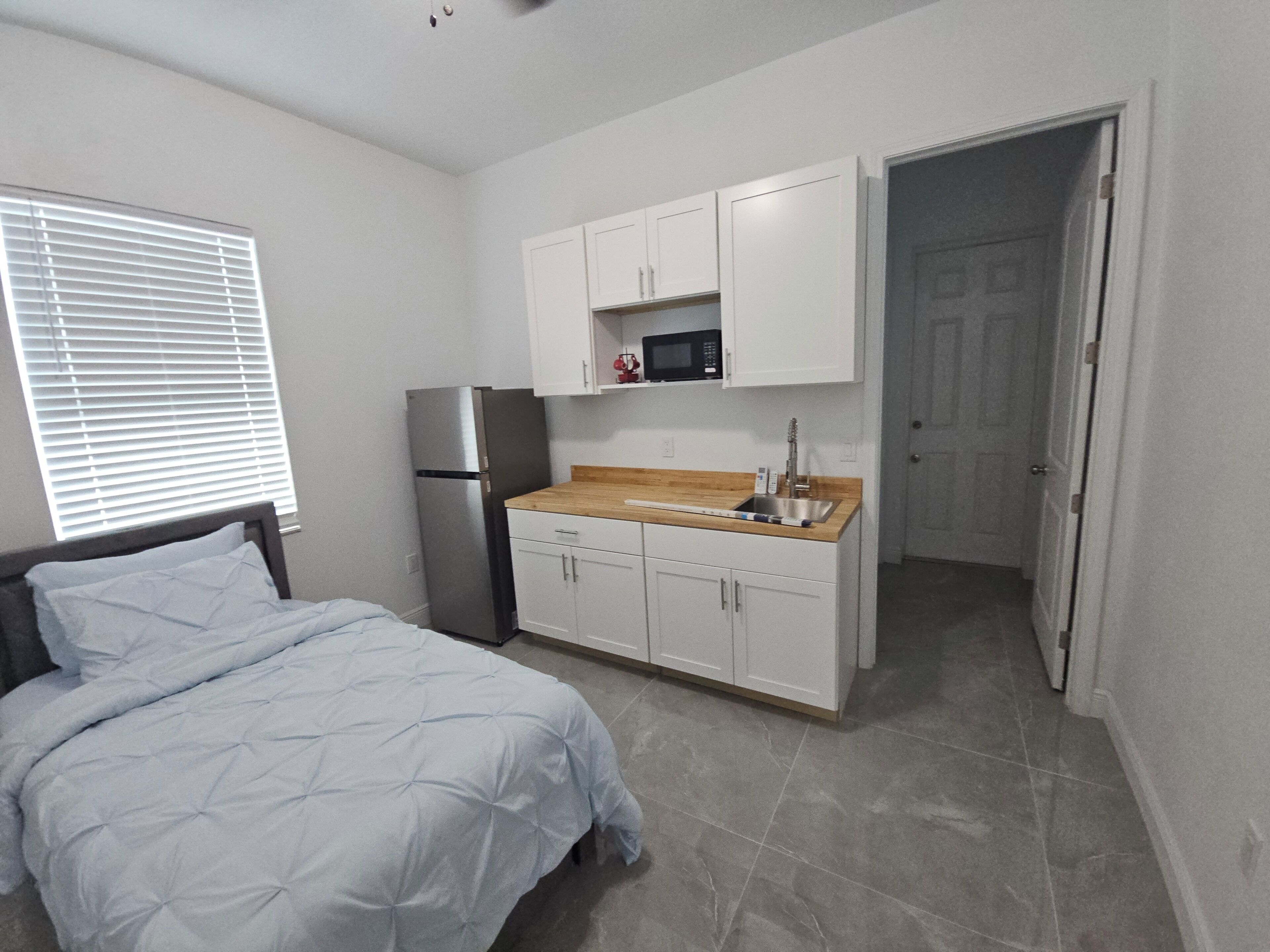 A compact bedroom with a bed, a built-in refrigerator, a microwave, and a sink in a kitchenette.