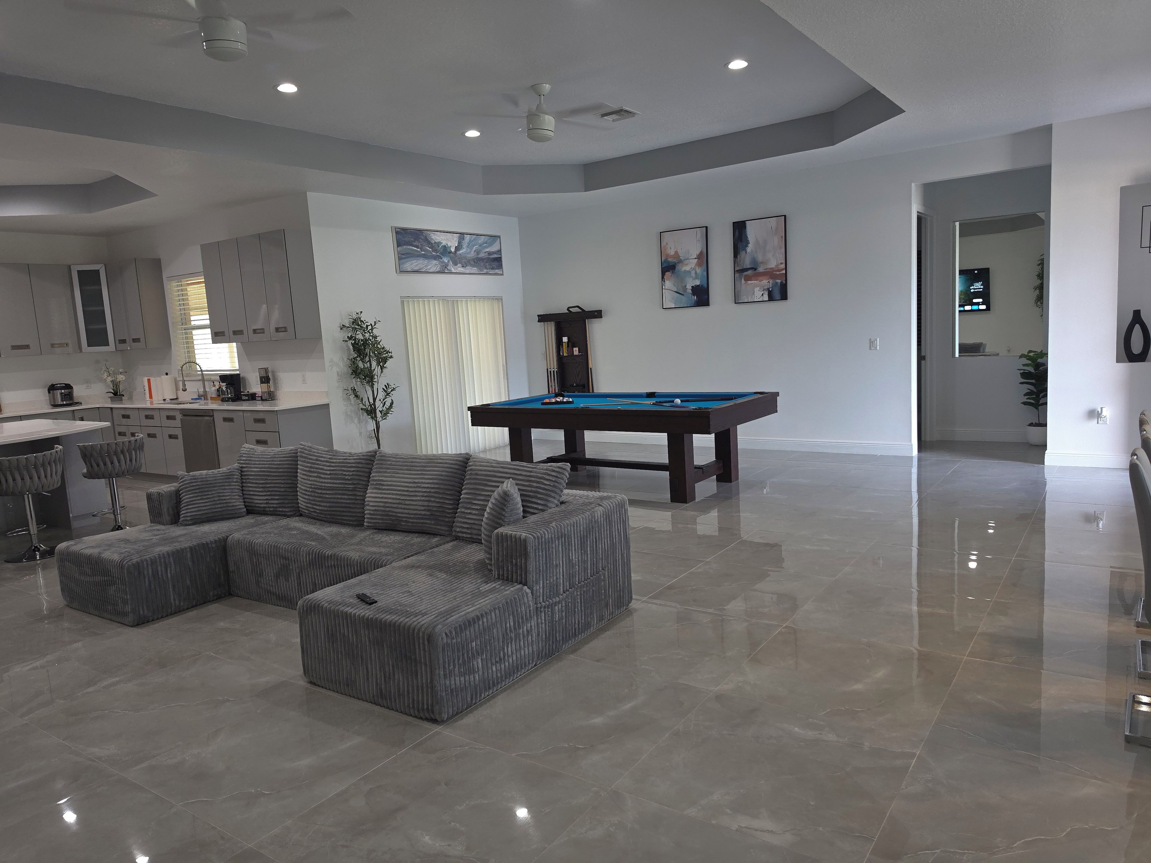 A modern living room with a pool table, grey sectional sofa, and a kitchen area with grey cabinets.