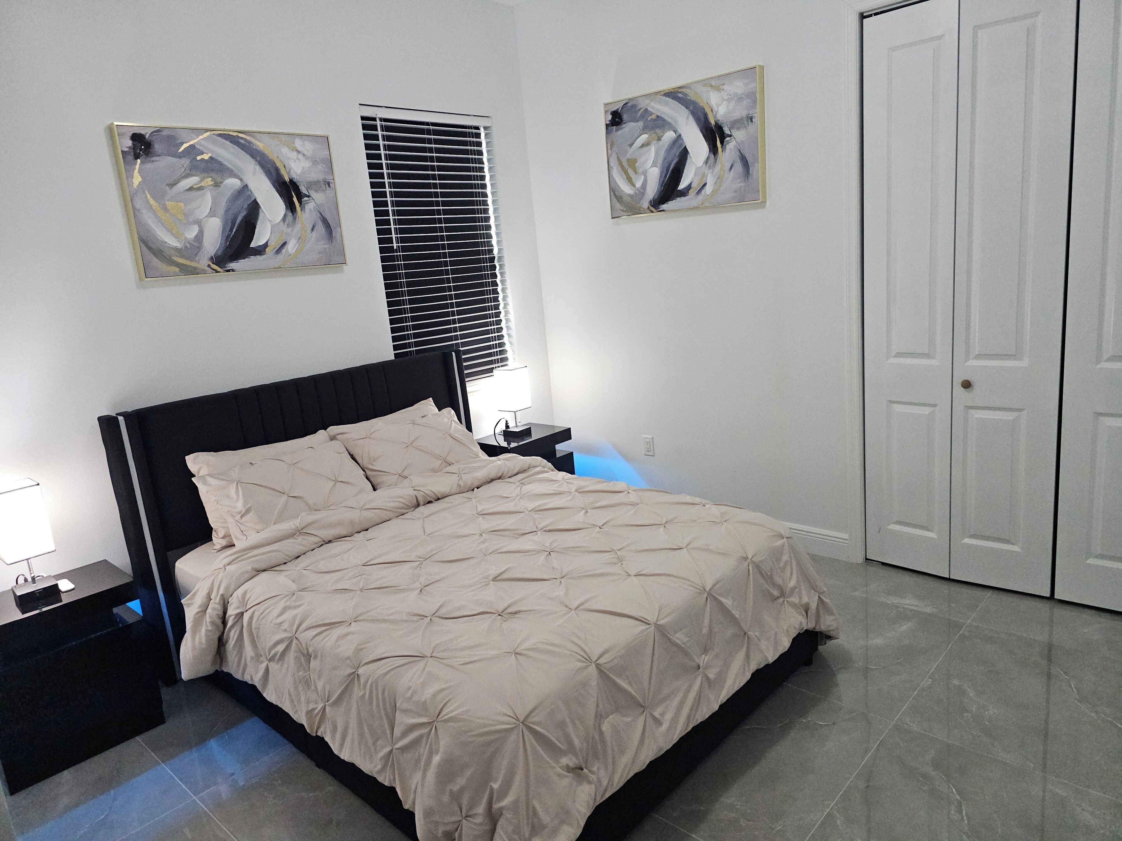Une chambre à coucher comprenant un lit, des tables de chevet, une commode, une fenêtre avec des stores et une porte.