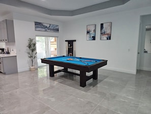 A modern living room with a pool table, a potted plant, and a kitchen area. - Modern  4-bedroom House  Very Spacious with Pool Table Game (Port St. Lucie)