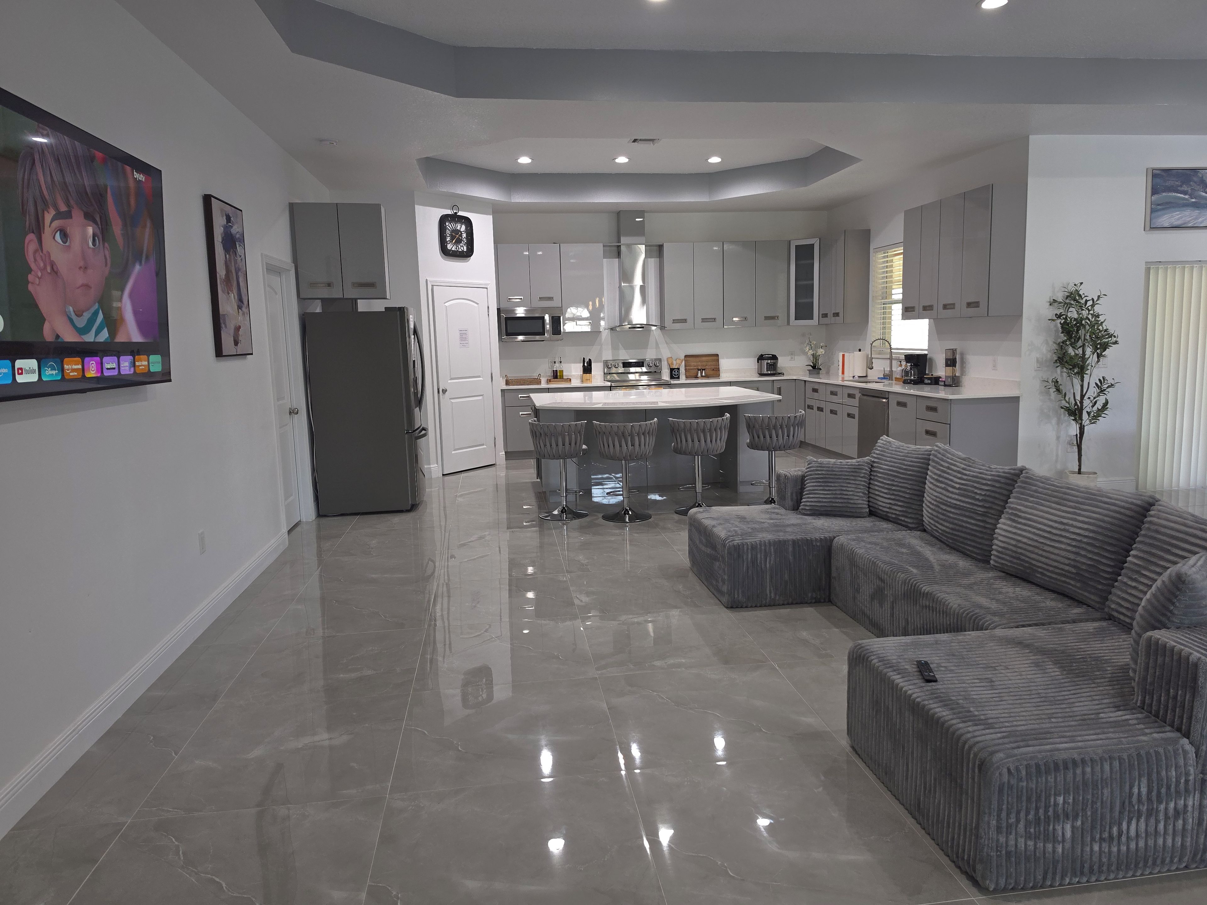 A modern kitchen with a central island, grey upholstered seating, and a flat-screen TV mounted on the wall.