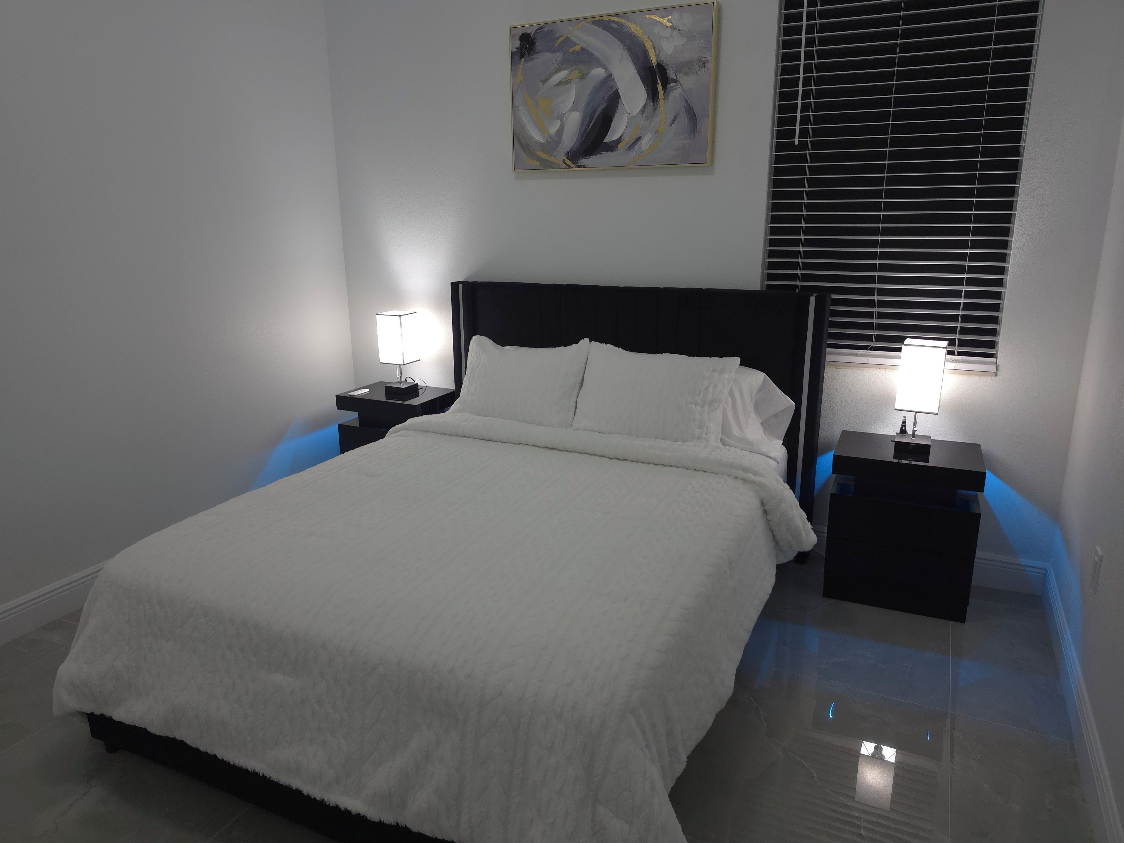 A neatly made bed with white bedding, two bedside tables with lamps, a wall art piece, and a window with blinds.