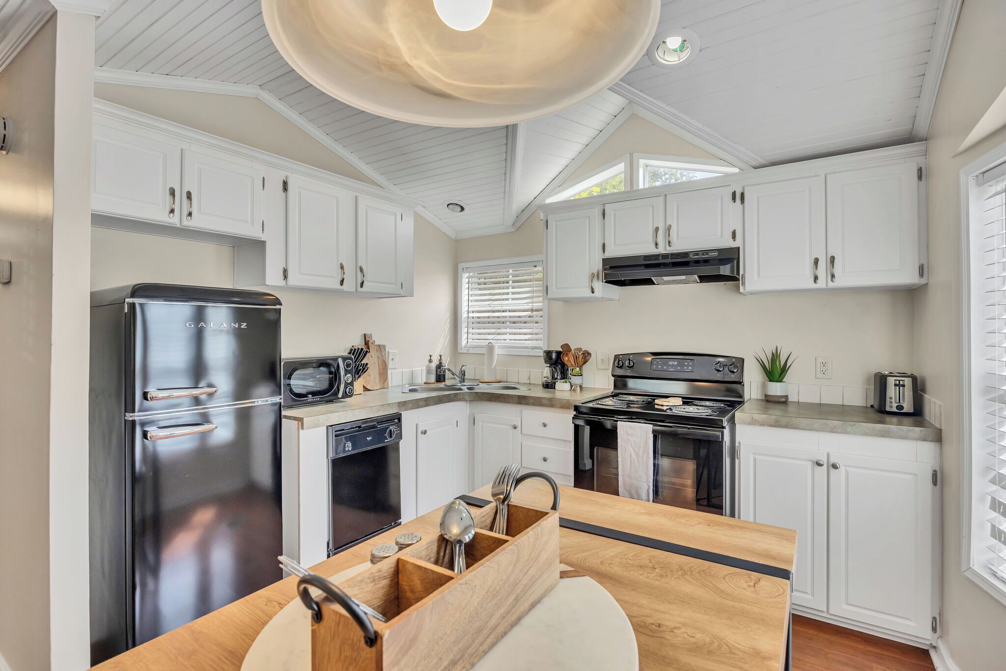 A modern kitchen with white cabinets, a stainless steel refrigerator, a dishwasher, a stove, and a wooden countertop with utensils.