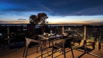 Terraza en la azotea con una mesa, sillas, plantas en macetas y vistas de la ciudad.