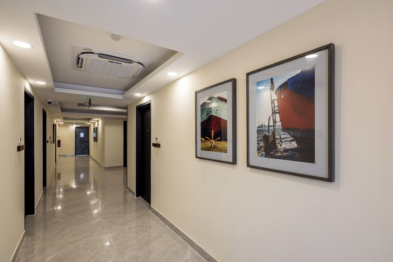 A hotel corridor with a polished floor, framed artwork on the walls, and a ceiling-mounted air conditioning unit.