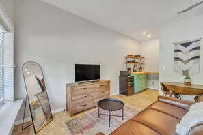 A modern living room with a flat-screen TV, a wooden dresser, a round coffee table, a sofa, and a kitchen area with a dining table and chairs. - Brand New, Adorable Suite Near the Caverns! (Monteagle)
