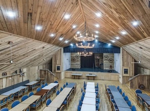 Un gran salón con paredes y techo de madera, que cuenta con un escenario con fondo decorativo, mesas y sillas dispuestas para un evento.