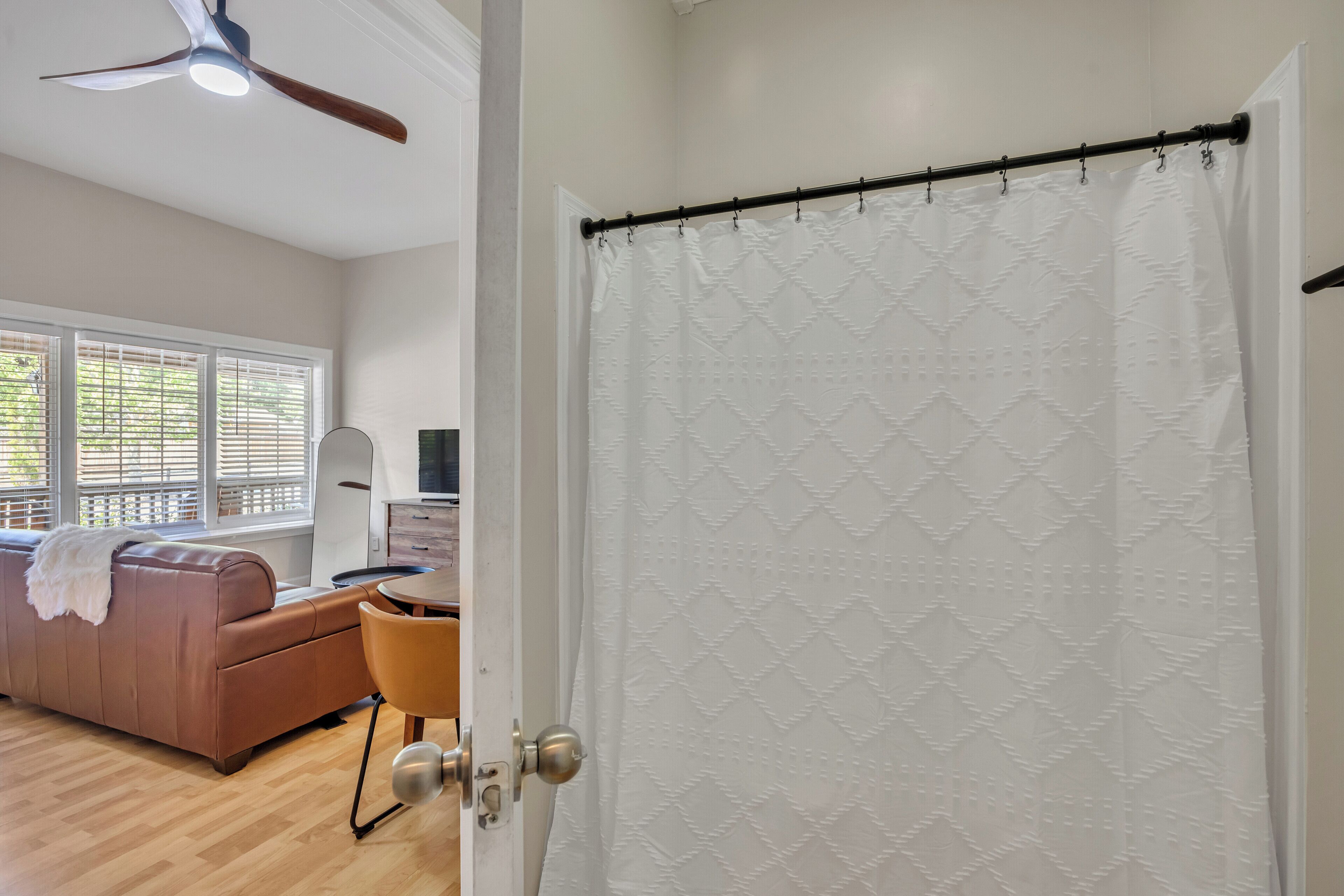 Une pièce avec un ventilateur de plafond en bois, une porte blanche ornée d’un rideau à motifs, un canapé marron et un plancher en bois.
