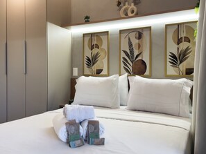 Una cama bien tendida con sábanas y almohadas blancas, un juego de toallas y una pequeña planta en maceta sobre la mesita de noche.