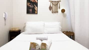 Ein ordentlich bezogenes Bett mit weißen Bettwäsche, ein an der Wand befestigtes Kunstwerk, ein Wandbehang aus Makramee und ein kleiner Nachttisch mit Lampe.