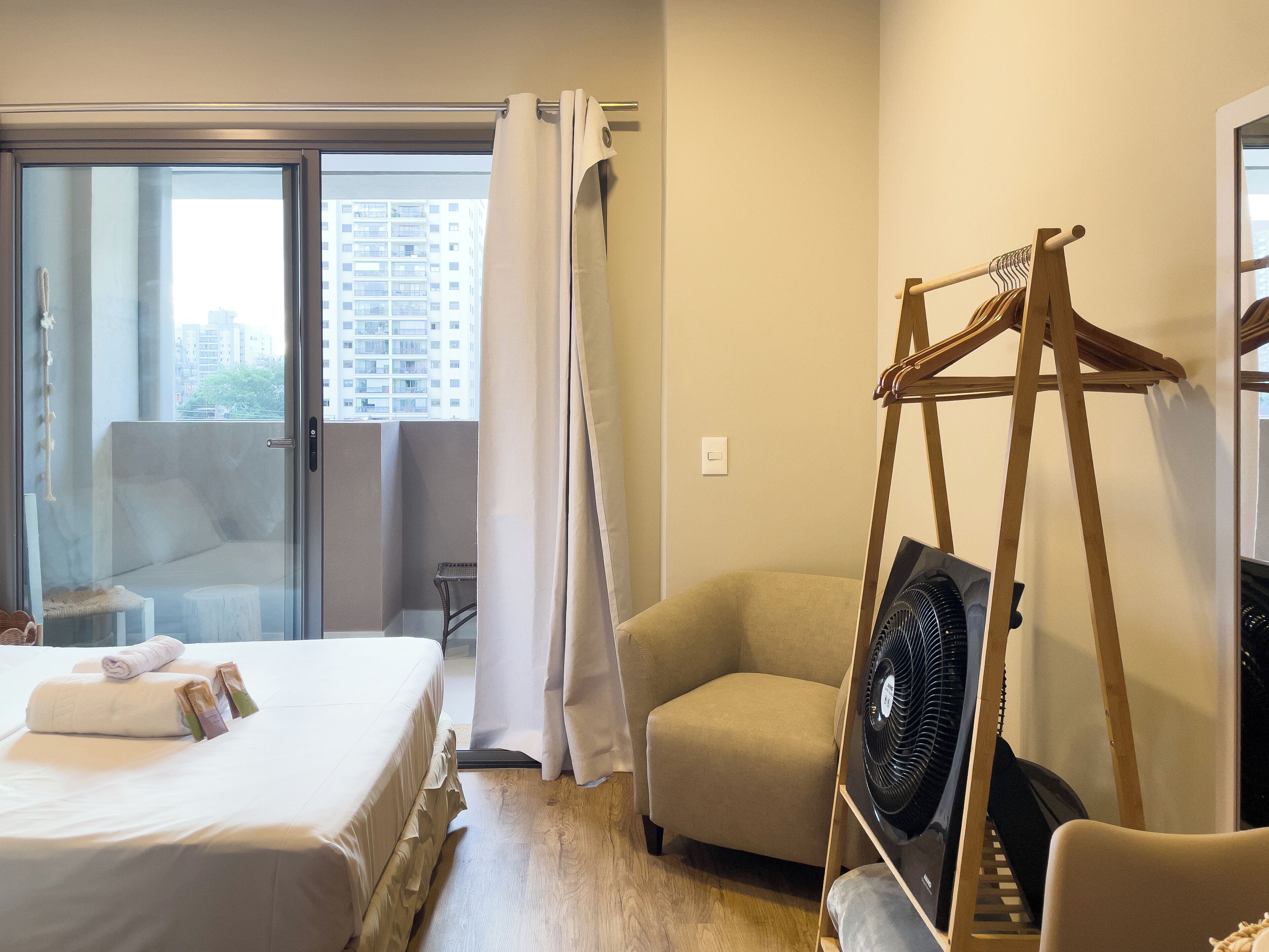 Habitació d'hotel amb un llit, una cadira, un ventilador i vista a la ciutat per la finestra.