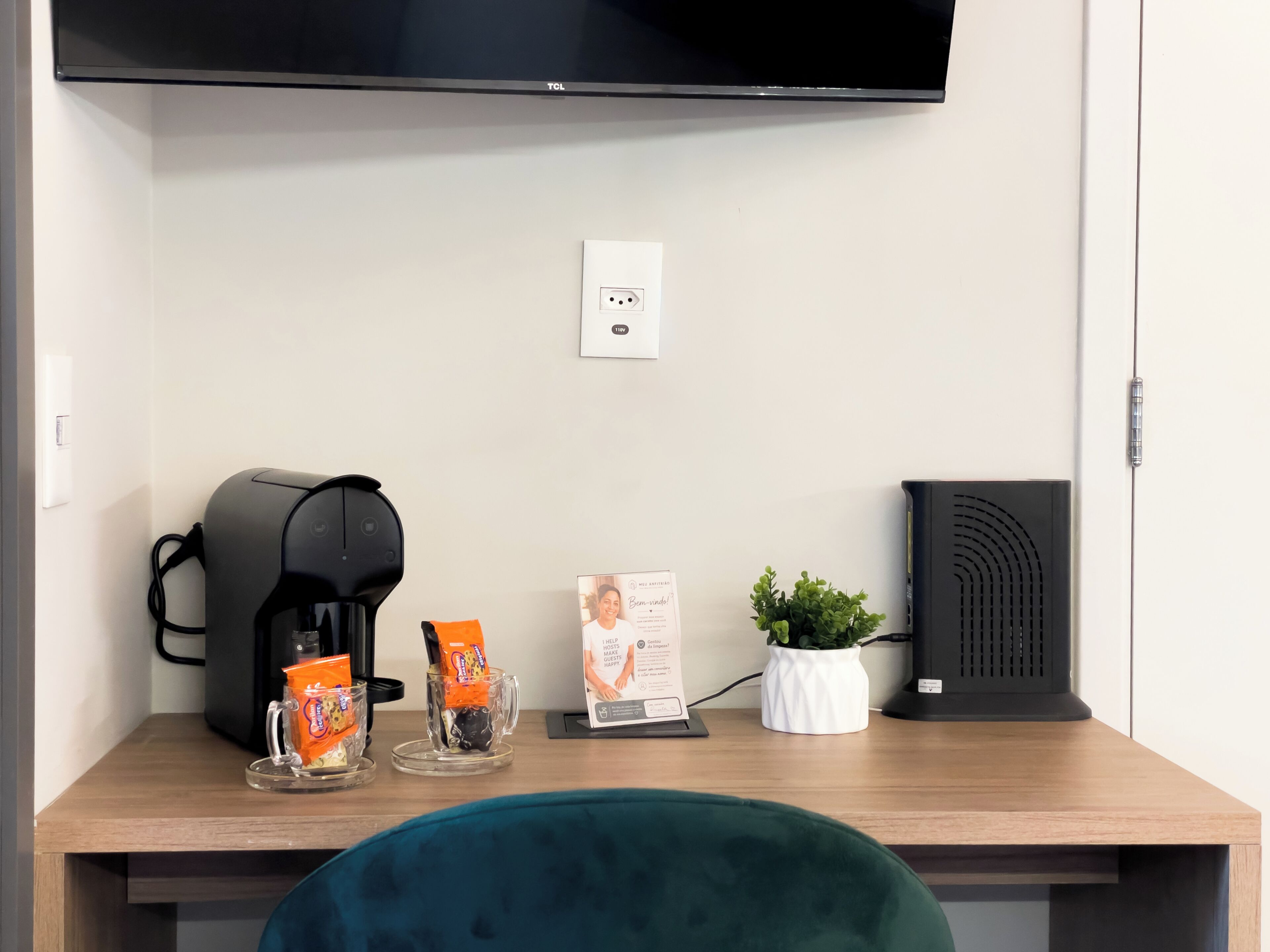 Habitación de hotel con un escritorio de madera, una cafetera negra, dos sobres de bebida anaranjada, una planta pequeña en maceta, un altavoz negro y un televisor de pantalla plana montado en la pared.