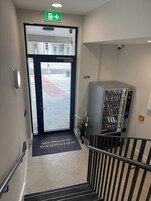 Entrada de un hotel con puerta de vidrio, un cartel de prohibido fumar, una máquina expendedora y un felpudo con el texto 'INTERNATIONAL AIRPORT'.