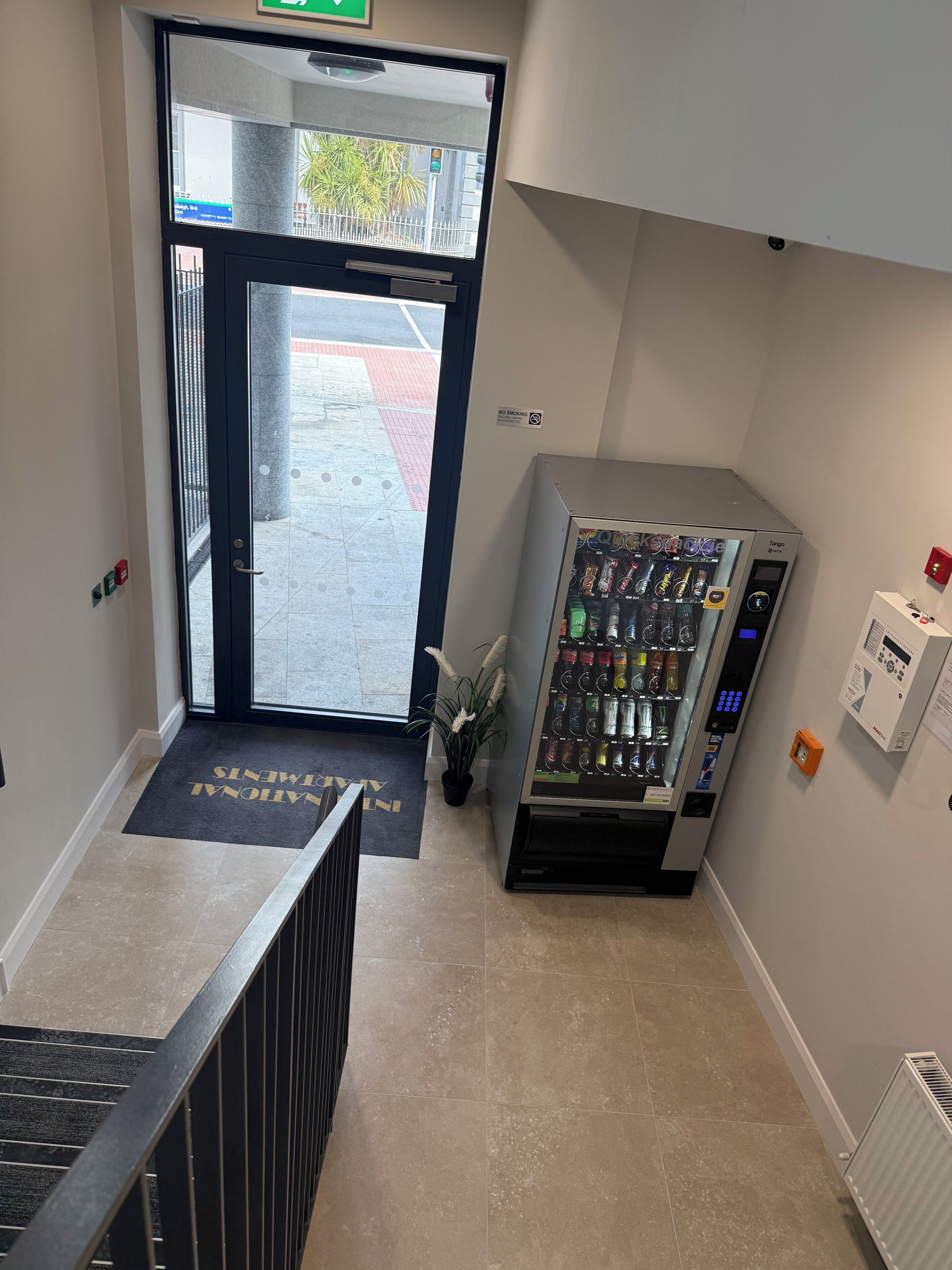 A hotel entrance with a vending machine, a 'No Smoking' sign, and a mat with 'Welcome' text.