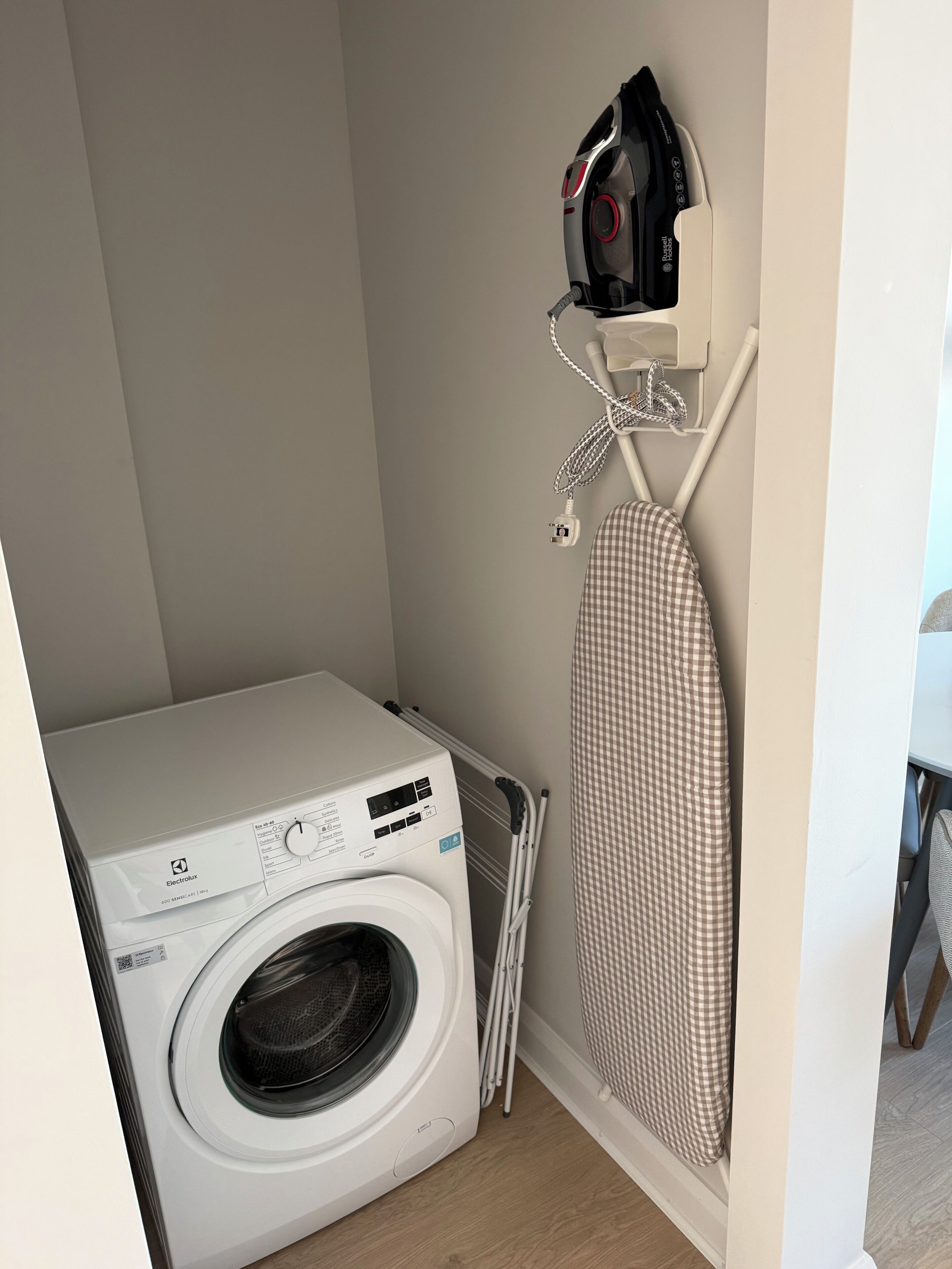 A compact laundry area with a front-loading washing machine, a drying rack, and a wall-mounted ironing board.