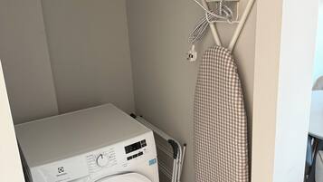 A compact laundry area with a front-loading washing machine, a drying rack, and a wall-mounted ironing board.