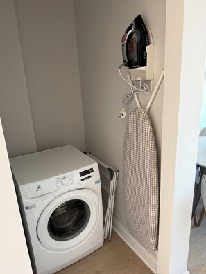 Un área de lavandería compacta con lavadora de carga frontal, un tendedero y una tabla de planchar fijada a la pared.