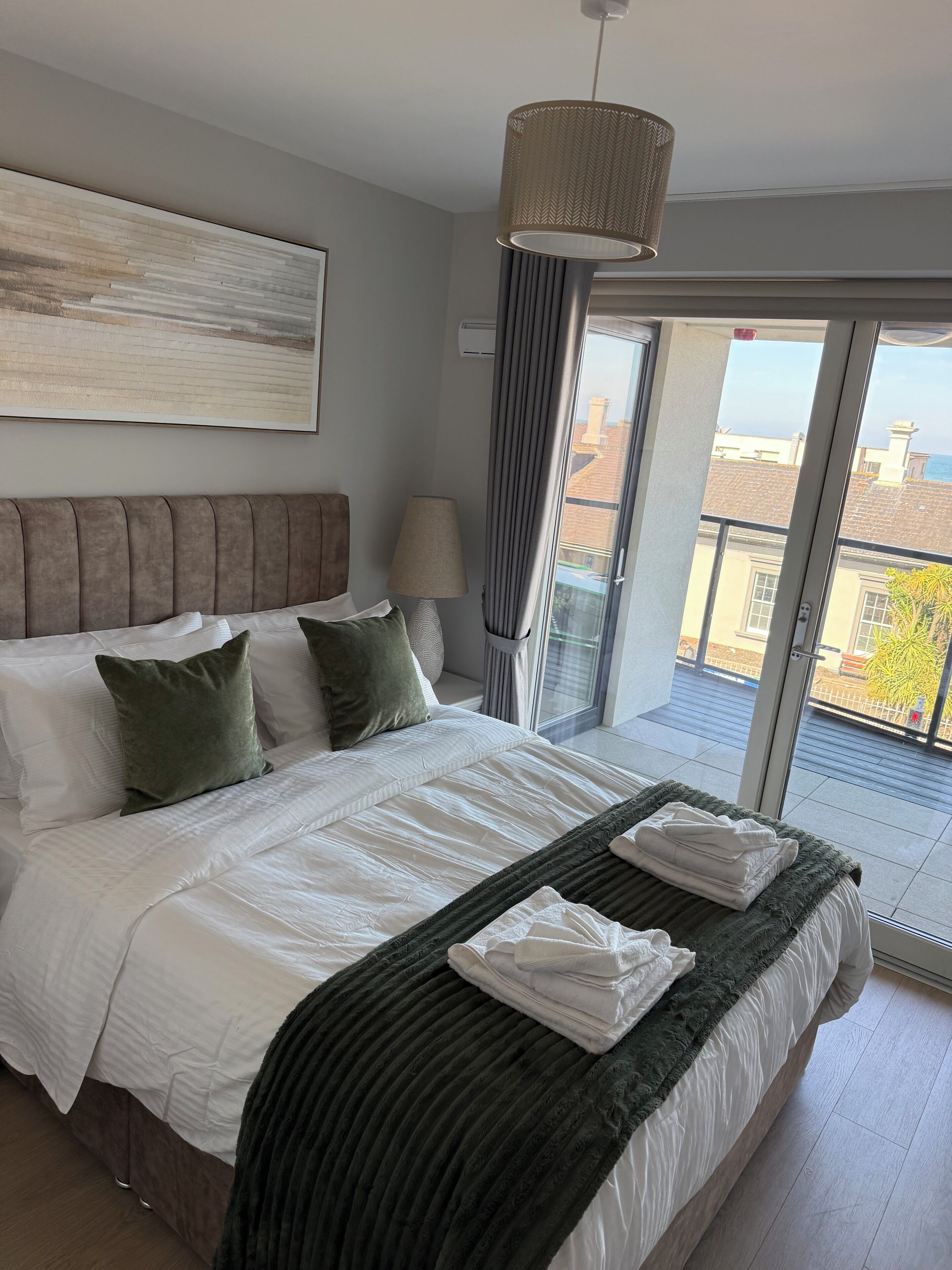 A modern bedroom with a large bed, a headboard, a bedside lamp, and a view of the outside through a sliding door.