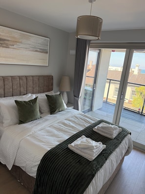 A modern bedroom with a large bed, a headboard, a bedside lamp, and a view of the outside through a sliding door. - The International Apartments Bray (Bray)