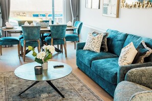 A living room with a teal sofa, matching chairs, a round coffee table, and a dining area with a table and chairs.