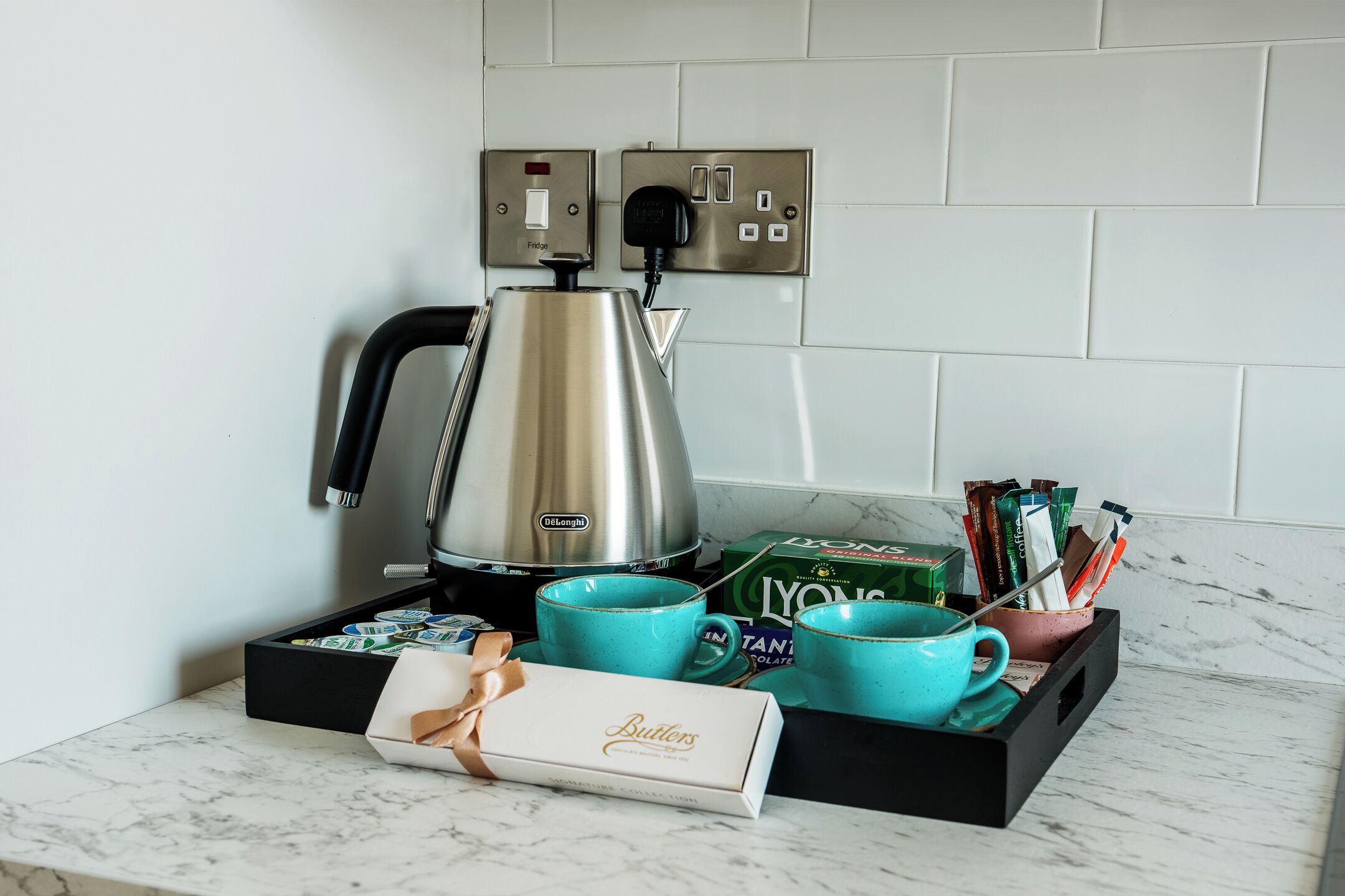 A coffee station with a De'Longhi espresso machine, two De'Longhi cups, a box of Butlers chocolate, and a box of L'Yonne coffee.