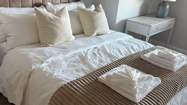 Una cama bien tendida con una colcha blanca y a rayas beige, una mesita de noche con una lámpara y un espejo montado en la pared.