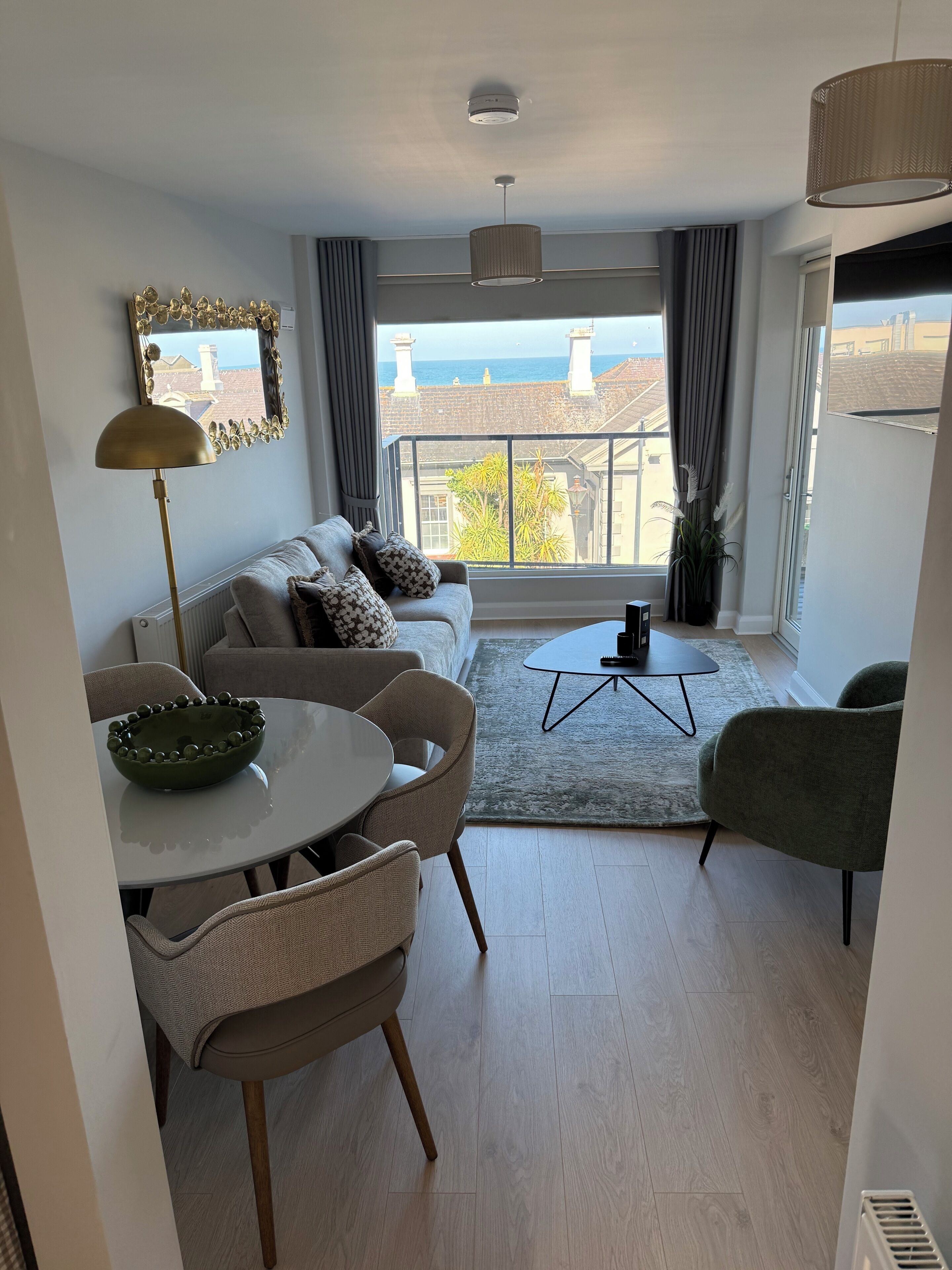 A modern living room with a sofa, dining table, chairs, a coffee table, and a view of the sea through large windows.