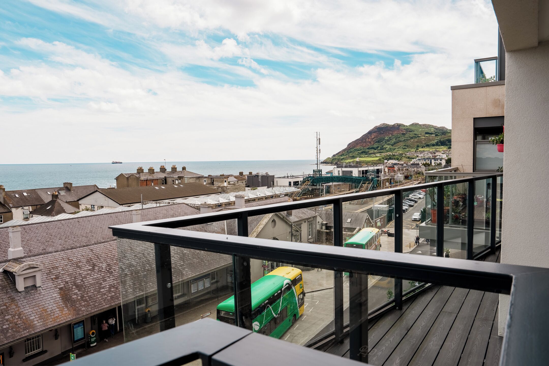 Een balkon met uitzicht op een kustplaatsje, een groene bus en een berg in de verte.
