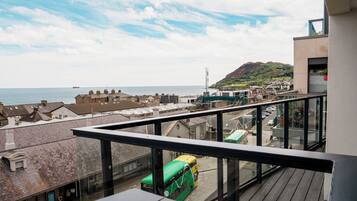 Een balkon met uitzicht op een kustplaatsje, een groene bus en een berg in de verte.