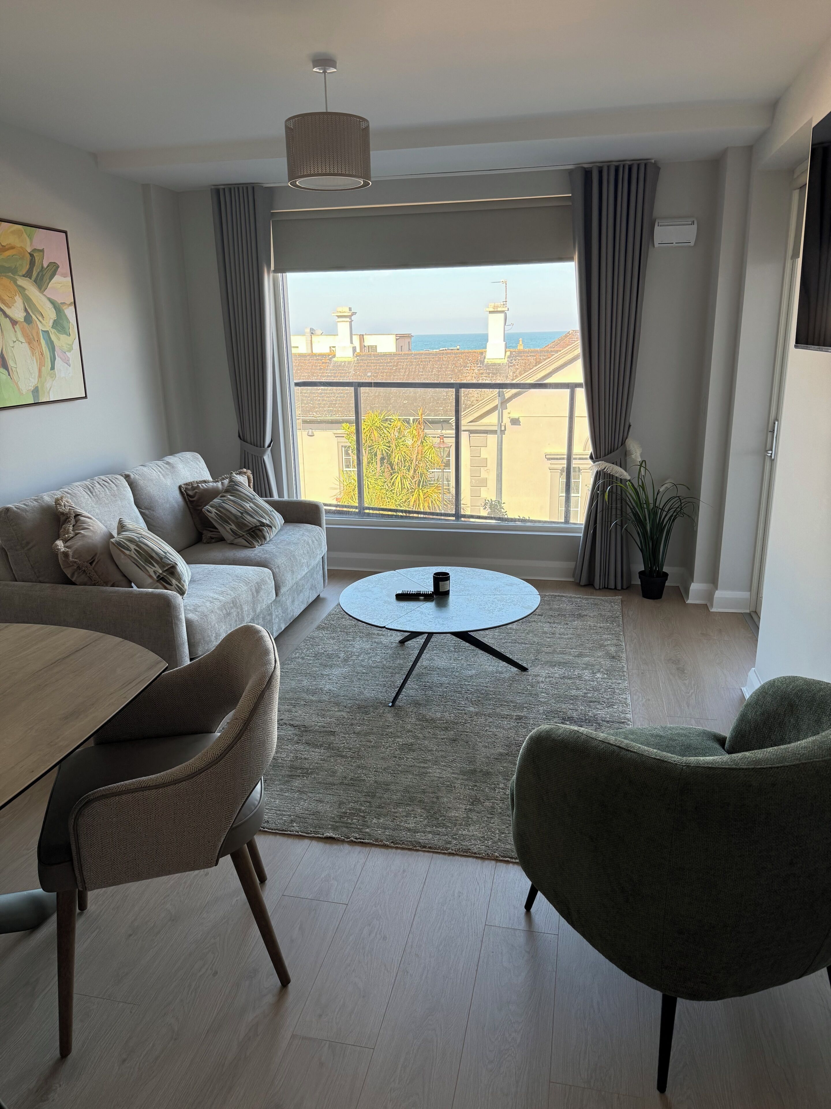 Uma sala de estar moderna com um sofá, cadeiras, uma mesa de centro e vista para o mar através de grandes janelas.