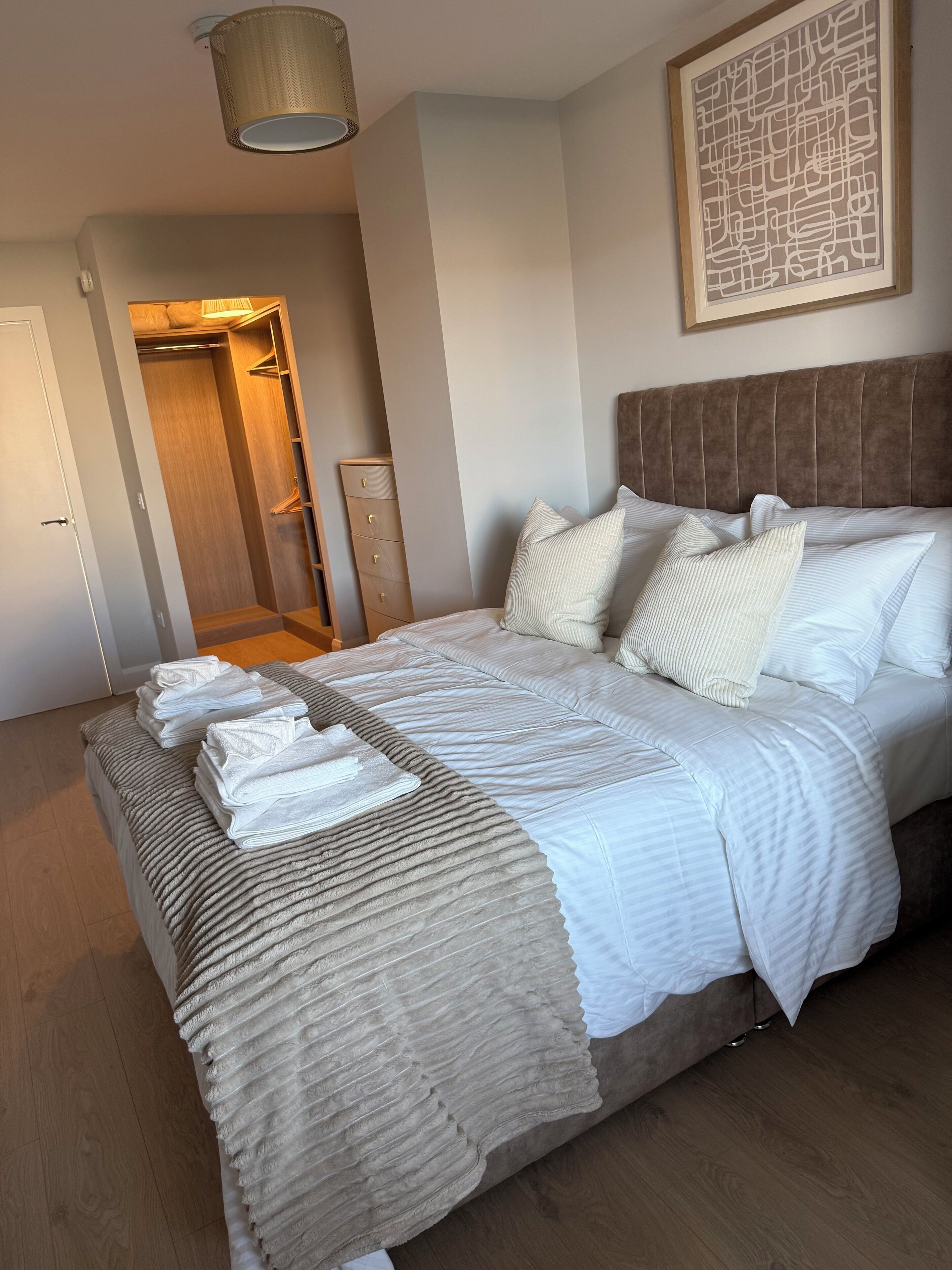 Un letto rifatto con cuscini e un asciugamano piegato, un armadio in legno, un quadro astratto incorniciato a parete e una porta che conduce in un'altra stanza.