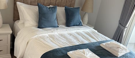 Uma cama bem arrumada com um cobertor azul e duas toalhas dobradas, uma cabeceira de madeira, uma mesa de cabeceira com uma luminária e um quadro abstrato grande na parede.