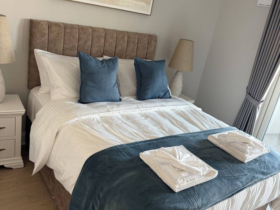 A neatly made bed with a blue blanket and two folded towels, a wooden headboard, a bedside table with a lamp, and a large abstract painting on the wall.