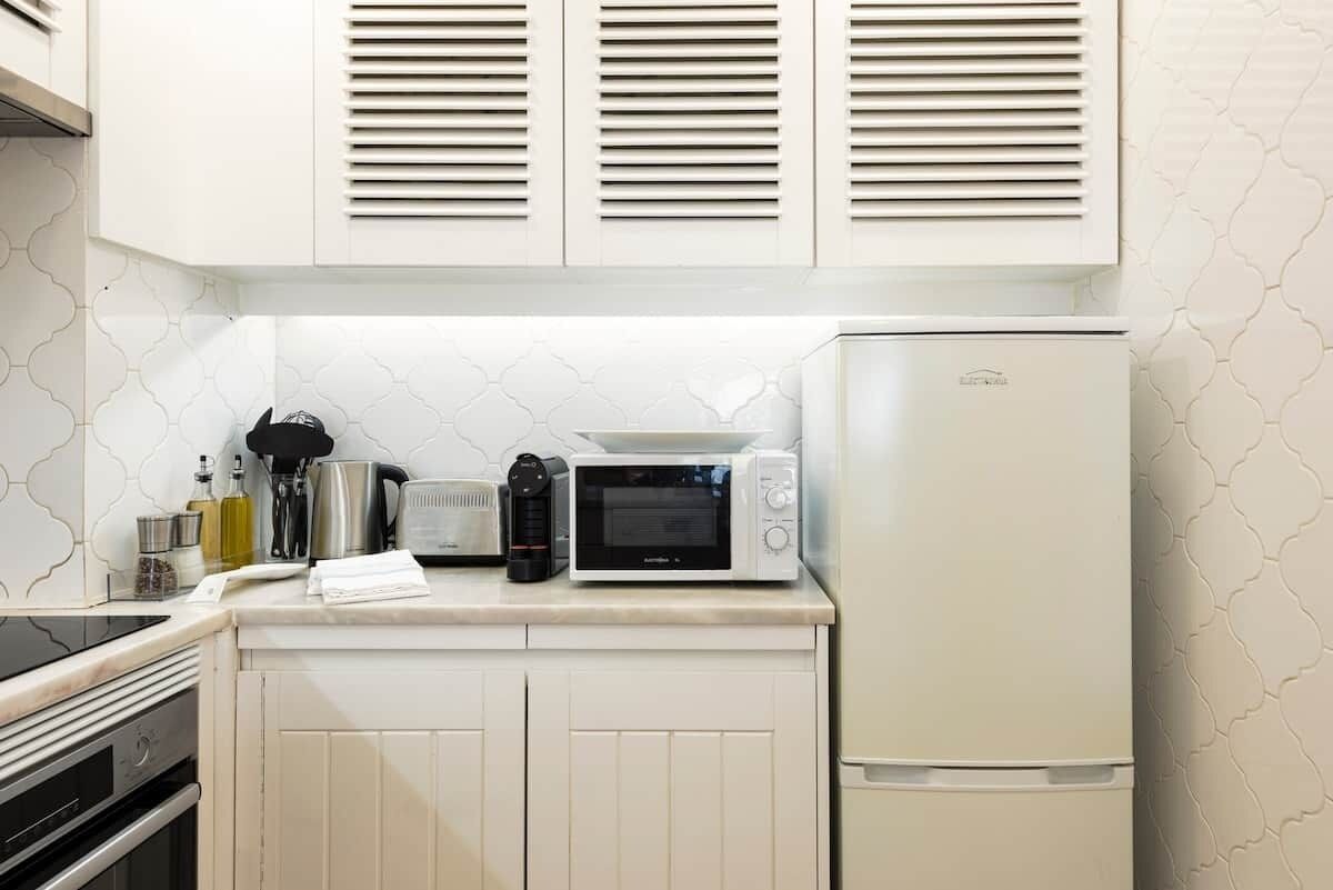 Una cocina con armarios blancos, microondas, hervidor y refrigerador.