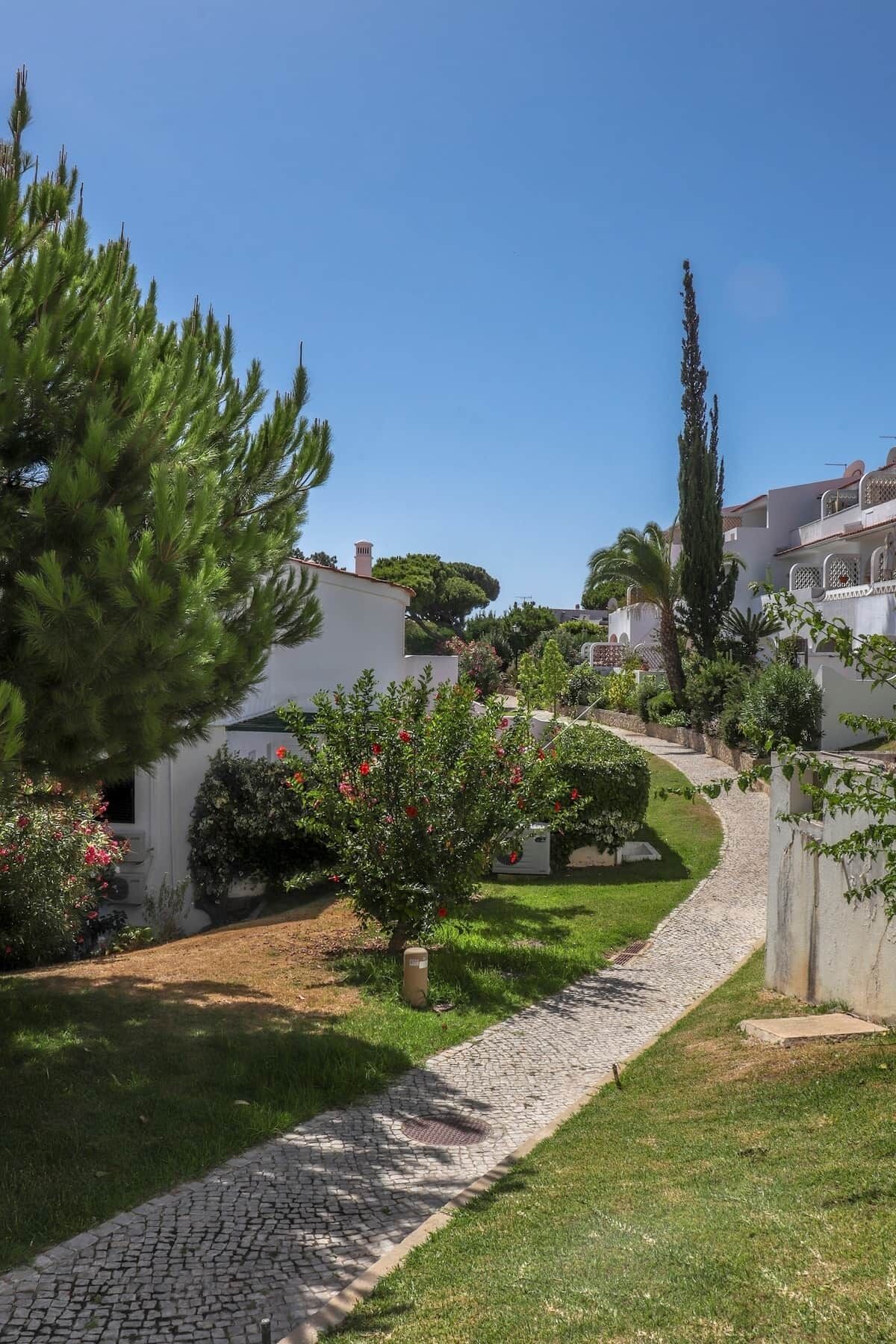A pathway leading through a garden with a variety of plants and trees, surrounded by buildings with white walls and red-tiled roofs.