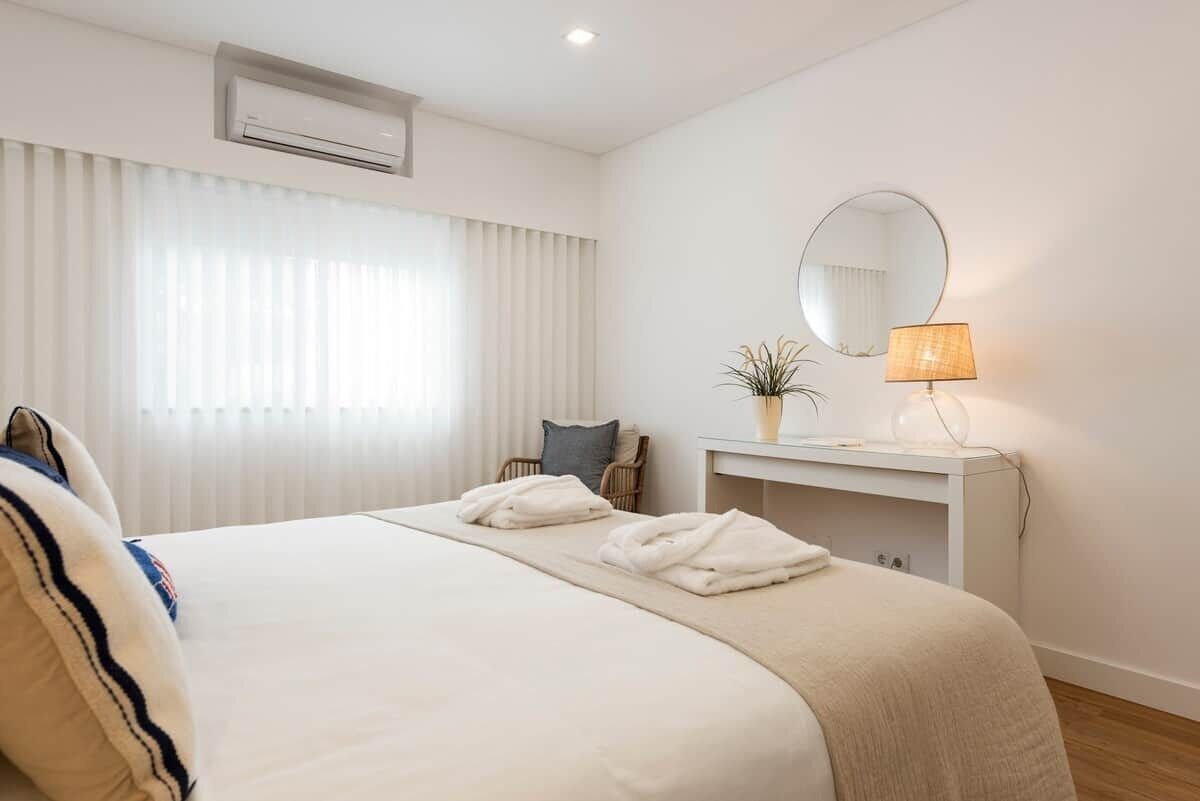 A hotel room with a bed, a bedside table with a lamp, a mirror, and a window with blinds.