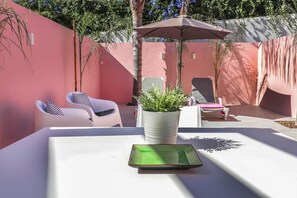 Eine sonnendurchflutete Terrasse mit einem weißen Tisch, zwei weißen Stühlen, einem Topfpflanzen und einem großen Sonnenschirm.
