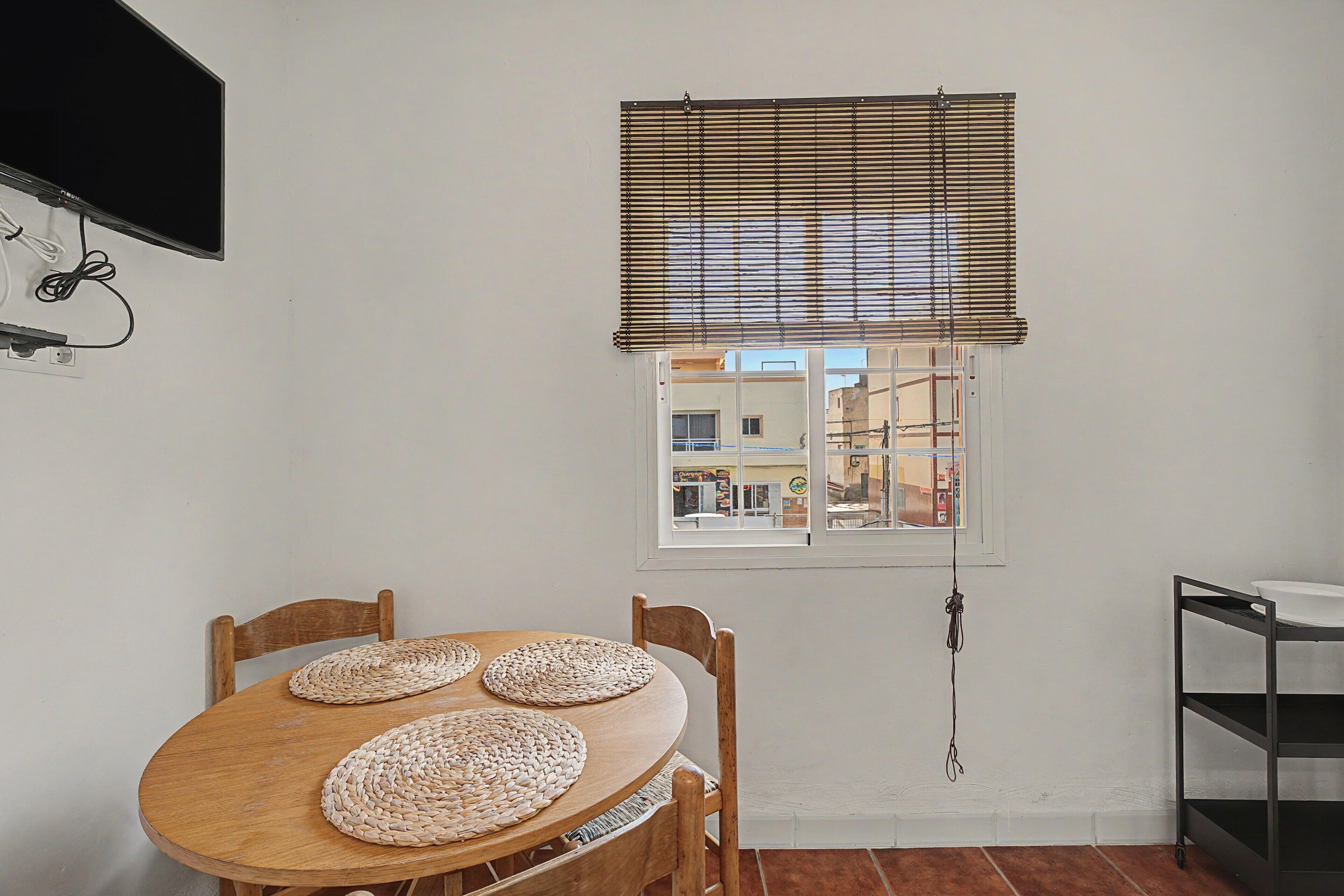 Un comedor con una mesa y sillas de madera, una ventana con persianas y un televisor montado en la pared.