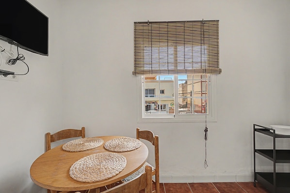 A dining area with a wooden table and chairs, a window with blinds, and a TV mounted on the wall.