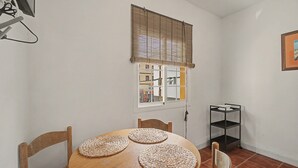 Una zona de menjador amb una taula rodona de fusta, quatre sotsplats teixits, dues cadires de fusta, una finestra amb persianes i un moble prestatge metàl·lic.