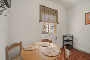 Uma área de refeições com uma mesa redonda de madeira, quatro individuais de palha, duas cadeiras de madeira, uma janela com persianas e uma estante de metal.