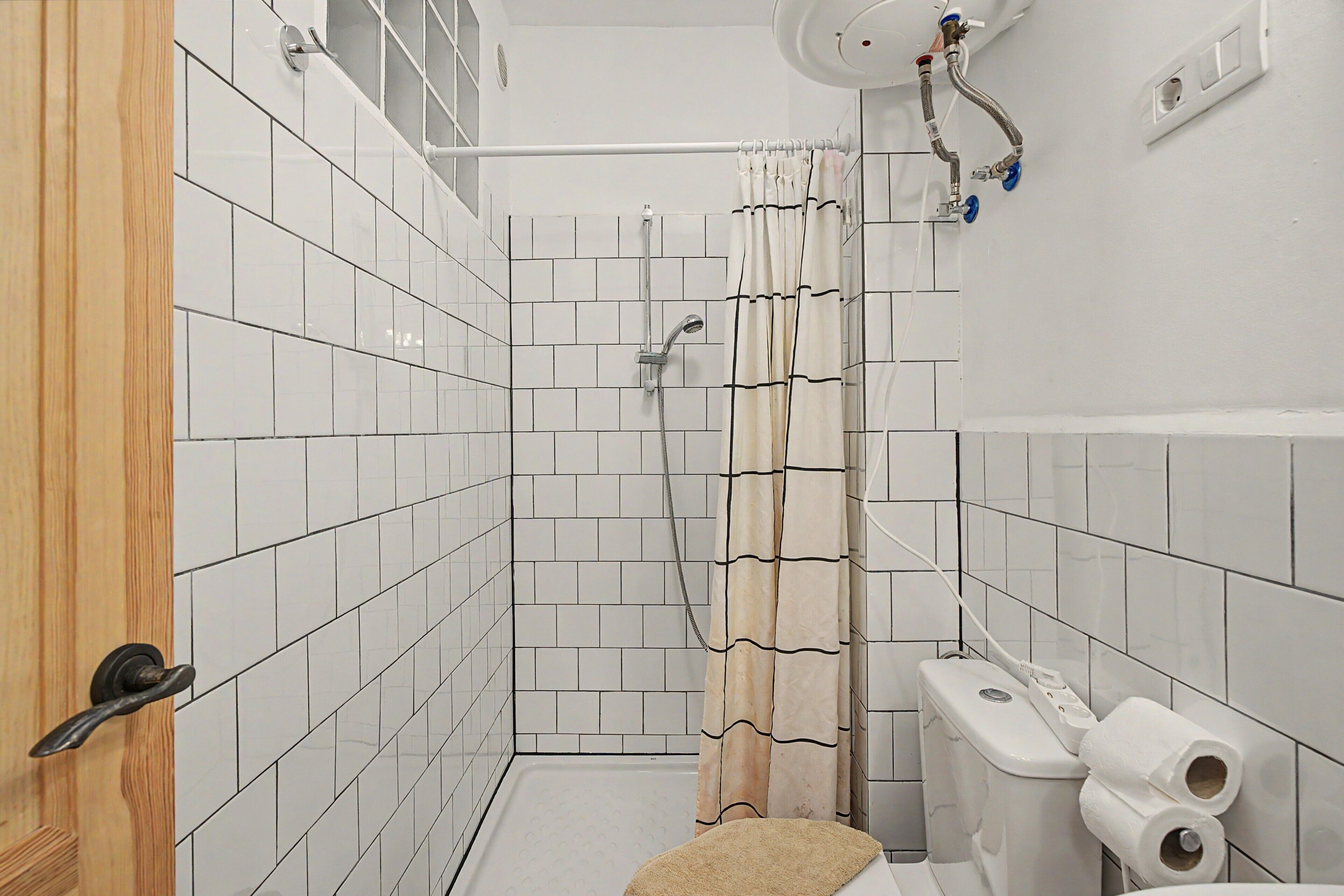 Une salle de bain avec des murs carrelés de carreaux blancs, une douche avec un rideau beige, des toilettes avec deux rouleaux de papier toilette et une porte en bois.