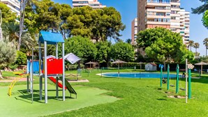 A playground with a slide and climbing structures, surrounded by grass and trees, with a pool in the background. - Cubo's Playamar Beach Apartment / communal pool / terrace / air conditioning / free Wi-Fi (Torremolinos)