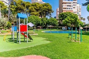 A playground with a slide and climbing structures, surrounded by grass and trees, with a pool in the background. - Cubo's Playamar Beach Apartment / communal pool / terrace / air conditioning / free Wi-Fi (Torremolinos)