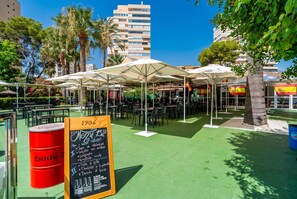 An outdoor café with tables and umbrellas, surrounded by greenery and buildings. - Cubo's Playamar Beach Apartment / communal pool / terrace / air conditioning / free Wi-Fi (Torremolinos)