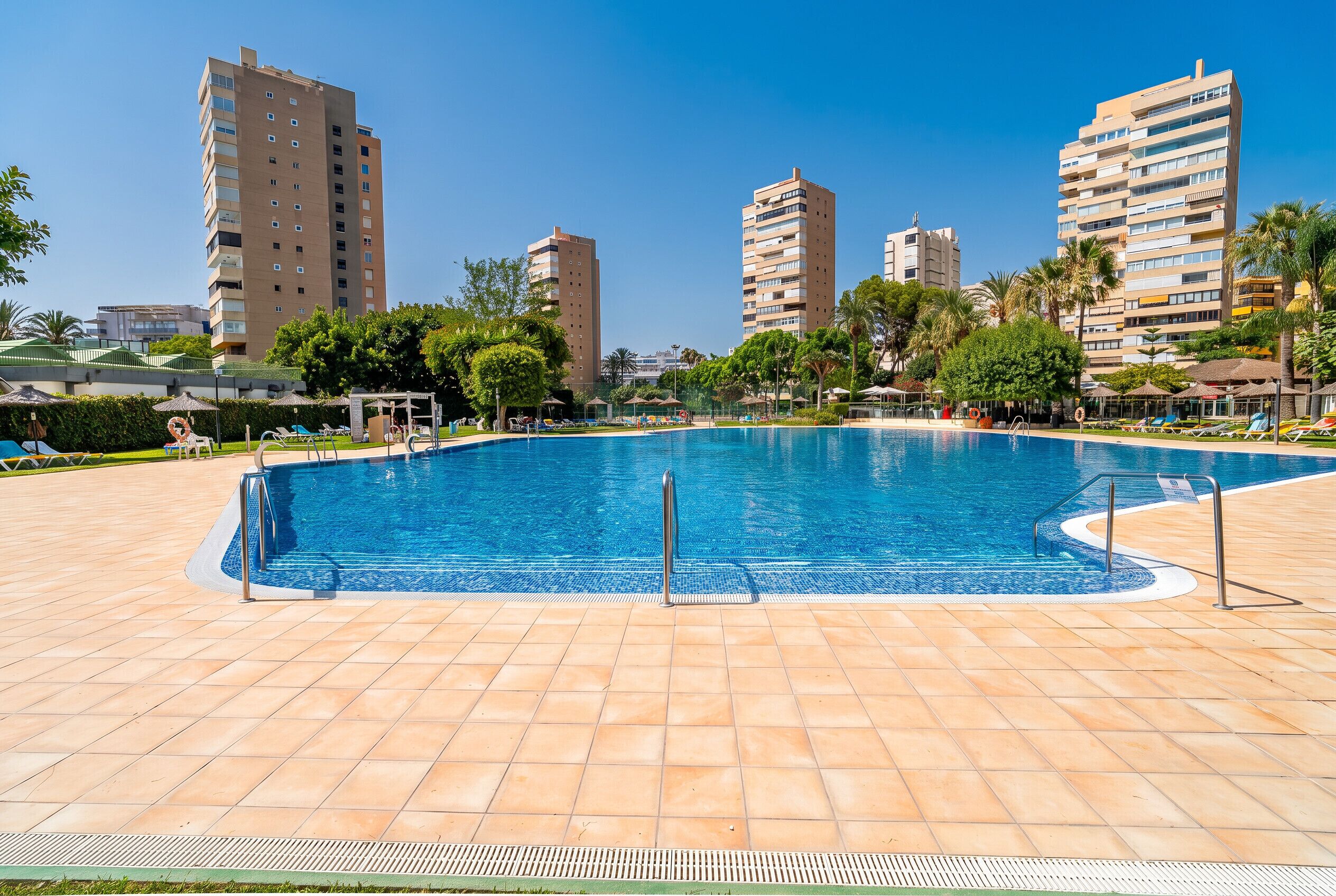 A swimming pool with a tiled deck, surrounded by palm trees and buildings.