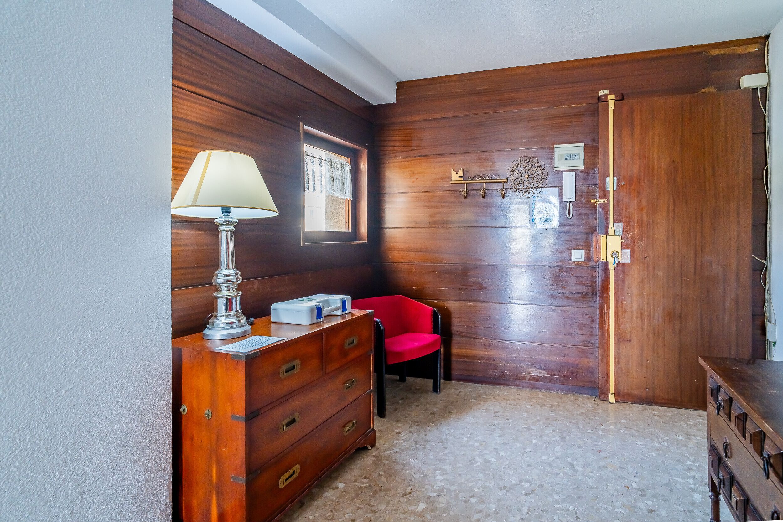 A room with a wooden wall, a lamp, a red chair, a wooden dresser, and a door with a key hook.