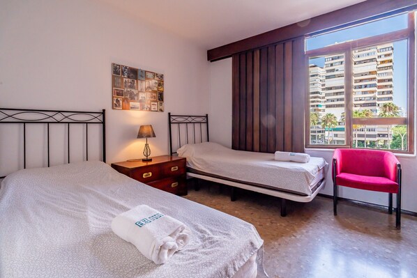 A hotel room with two beds, a nightstand, a lamp, a chair, and a window with a view of buildings. - Cubo's Playamar Beach Apartment / communal pool / terrace / air conditioning / free Wi-Fi (Torremolinos)